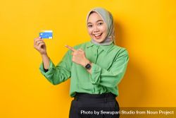 Muslim woman in headscarf and green blouse holding credit card smiling while pointing to it 42Aa35