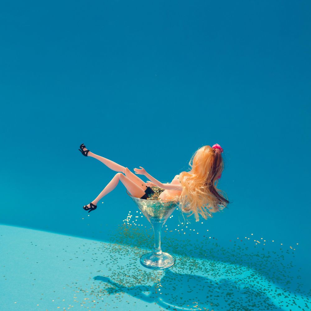 Doll bathing in martini glass full of gold glitter on blue background