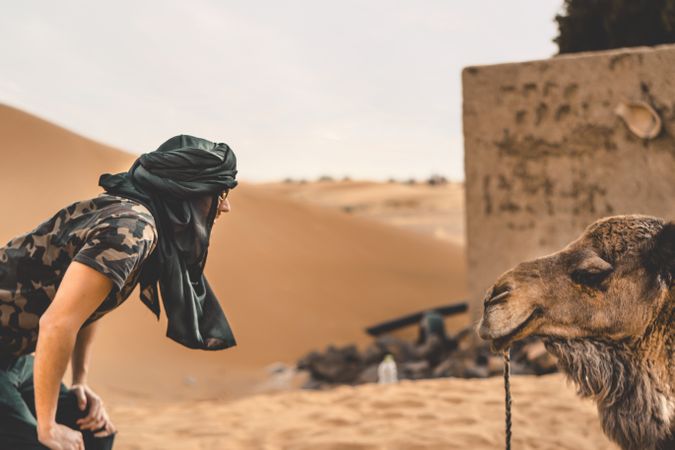 Man with dark headwear looking at a camel in desert
