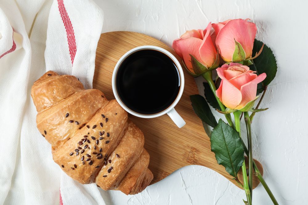 Composition with croissant, cup of coffee, roses and kitchen towel on light background, top view