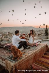 Man and woman raising toast on rooftop with hot air balloons in sky 0LZzP4