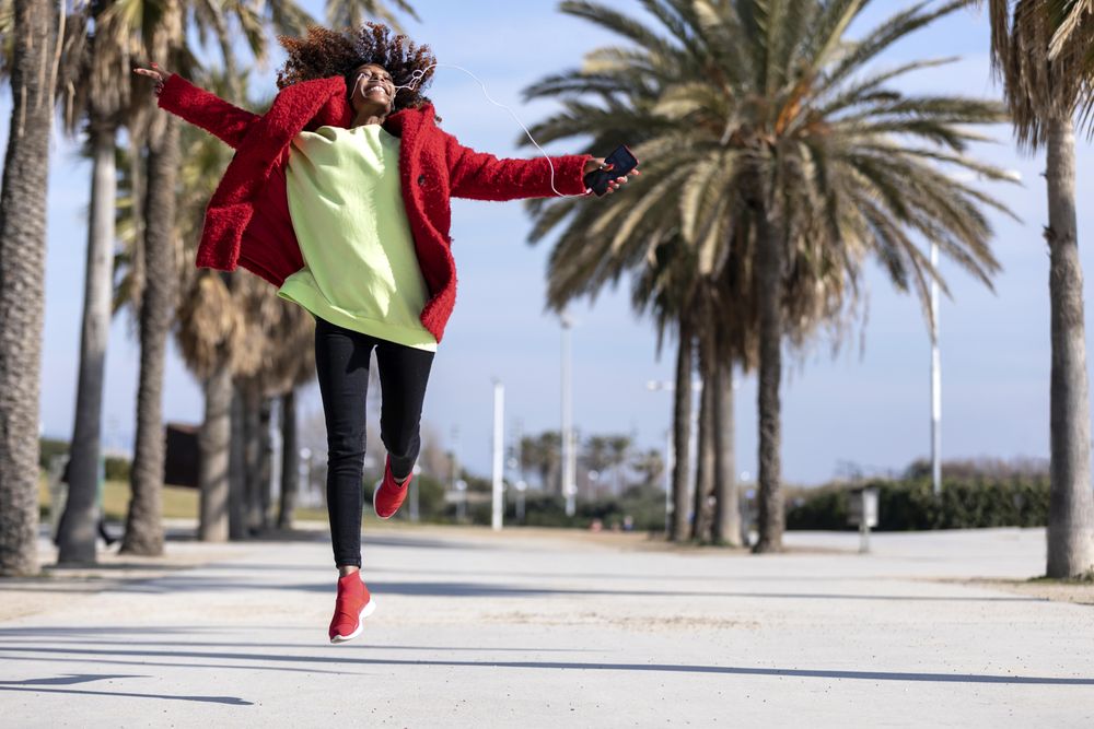Female jumping with open arms while listening to music outside on street with palm trees
