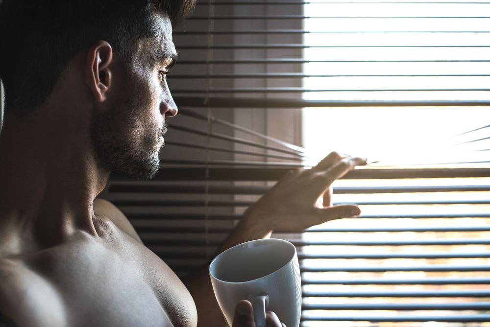 Man standing by a windows blind with a cup of coffee