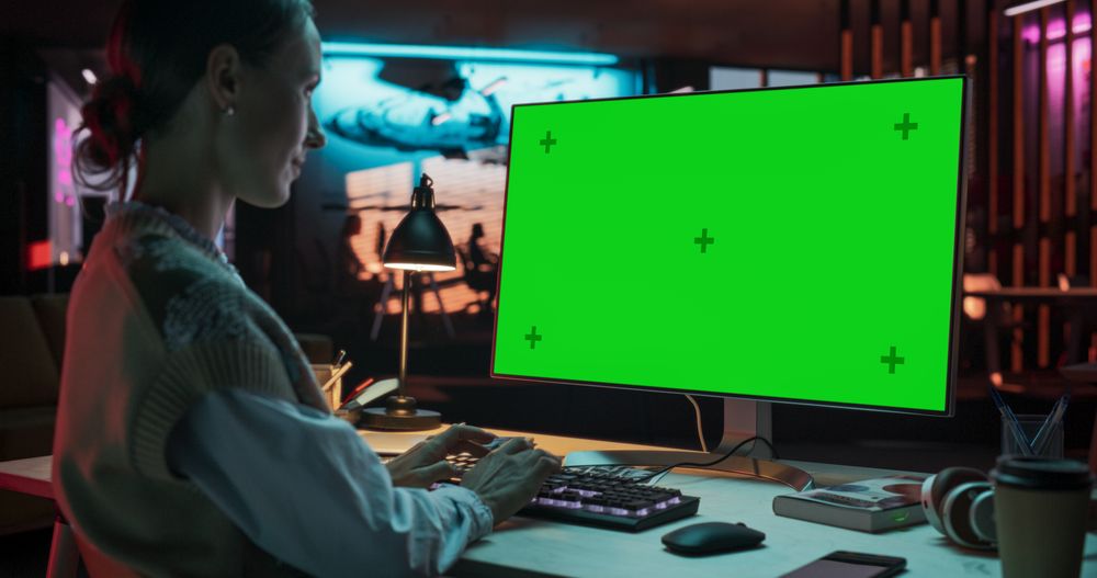 Over the Shoulder: Young Woman Sitting at Her Desk Using Desktop Computer with Mock-up Green Screen.