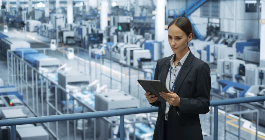 Thoughtful Female Engineer Monitoring and Analyzing Conditions at a Modern Tech Electronics Factory with Automated Robot Arms Working with the Help of Tablet Computer