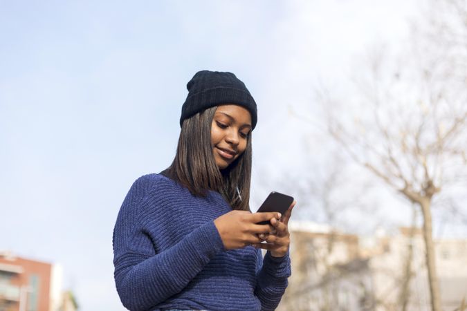 Female in wool hat and blue sweater checking smart phone on sunny day with space for text