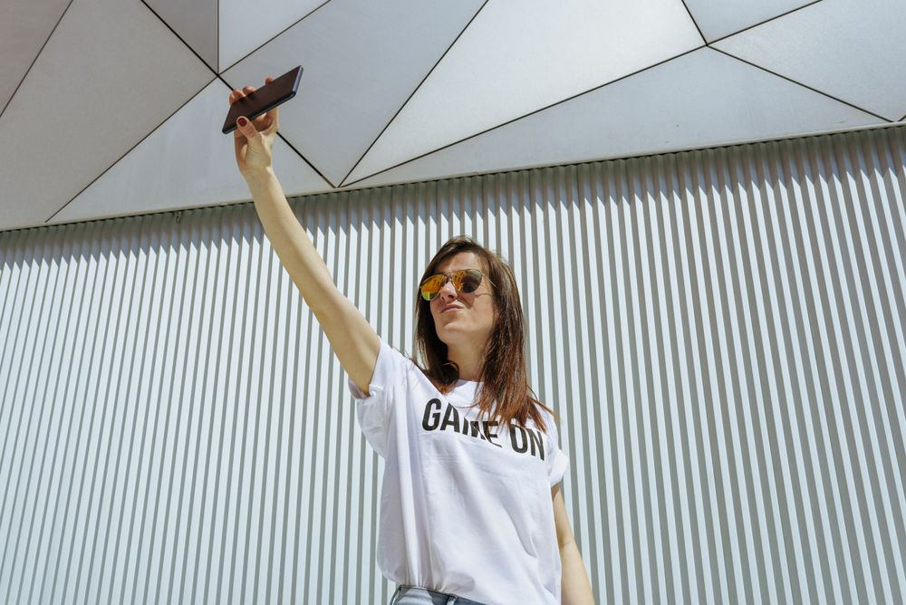 Cheerful woman taking selfie and posing near modern wall