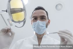 Dentist wearing surgical mask while holding angled mirror and drill, ready to begin 43XVrb