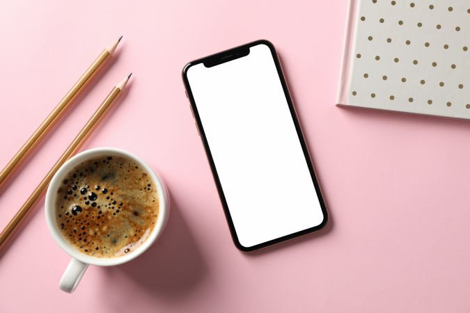 Phone with empty screen, copybook, pencils and cup of coffee on pink background, top view