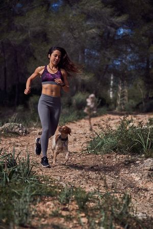 Active young Hispanic sportswoman with dog running along rough p
