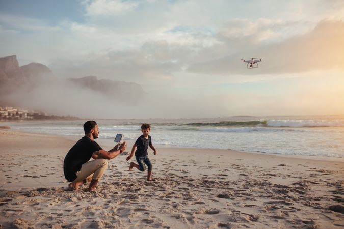 Father flying drone while son runs on sandy beach