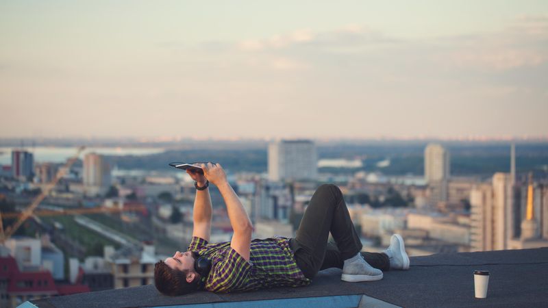 Male lying back with tablet, headphones and takeaway coffee cup on rooftop