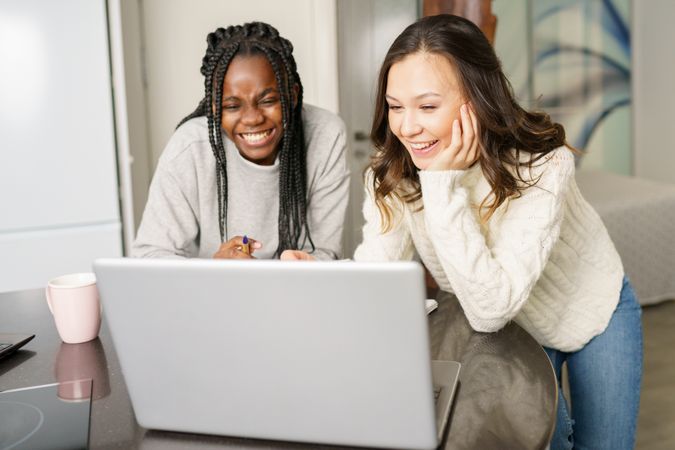 Two women sitting at kitchen table and laughing at something on a laptop