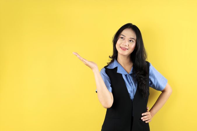 Asian Woman Wearing Blue Shirt Pointing to an Empty Space in a Studio with Yellow Background.