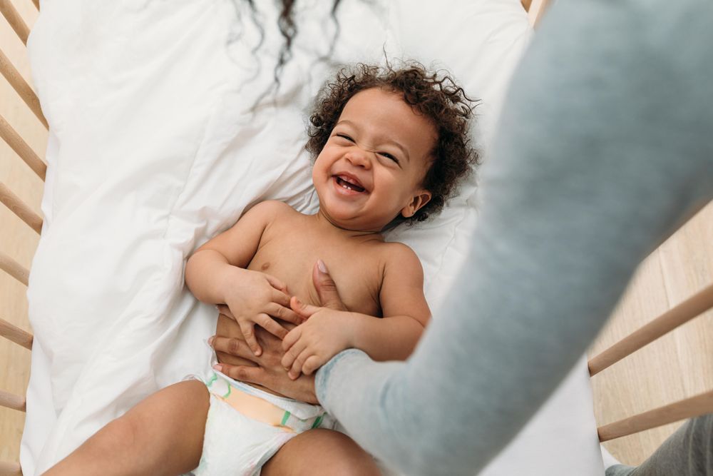 Happy baby boy lying in his crib with his mother’s arm tickling his belly