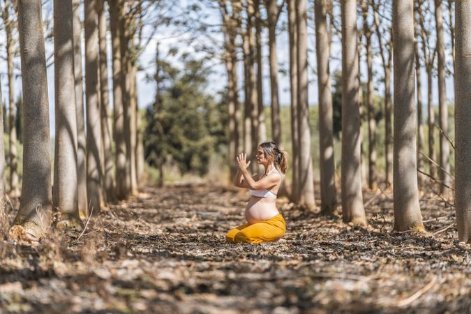 Calm adult pregnant woman practicing yoga while sitting in pose lotus on ground in park during sunny day