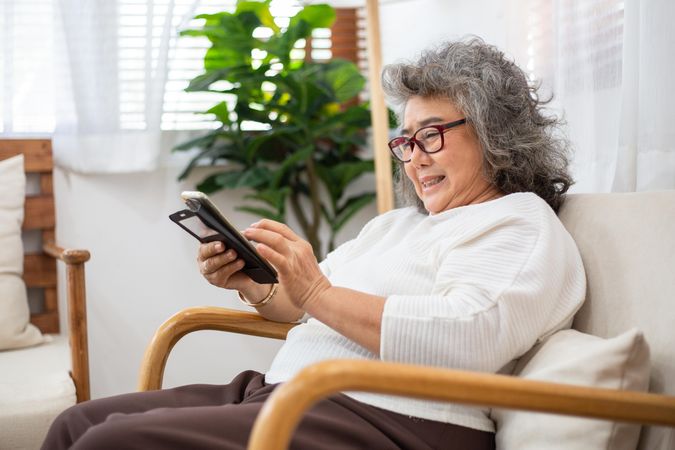 Happy older woman smiling and sitting on couch using smartphone