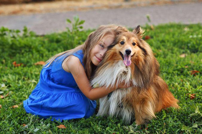 Girl in blue dress with flower in hair hugging her pet dog in the grass