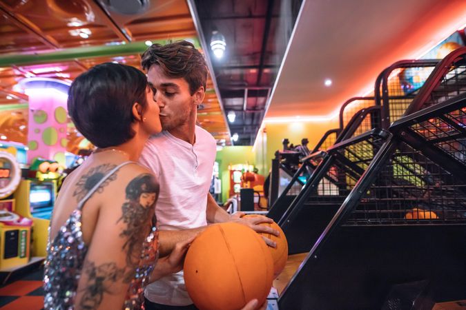 Couple kissing each other standing in a gaming parlour holding basketballs