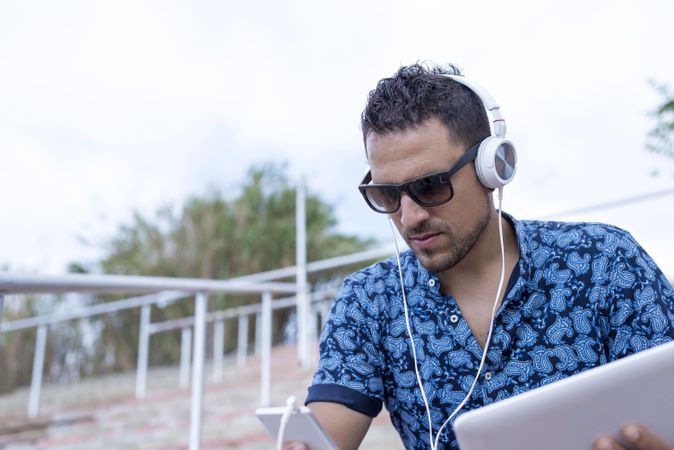 Man in sunglasses sitting outside with headphones, tablet and smartphone