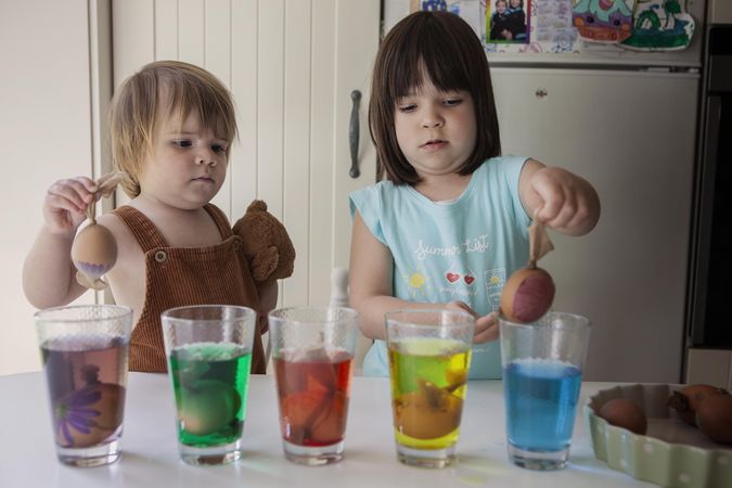 Girl and toddler putting eggs in coloring glasses in the kitchen at home