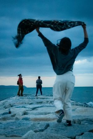 Back view of woman holding a scarf standing near people on rocky seashore