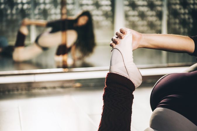 Crop shot of ballerina pulling leg to stretch muscles sitting on floor in ballet studio.
