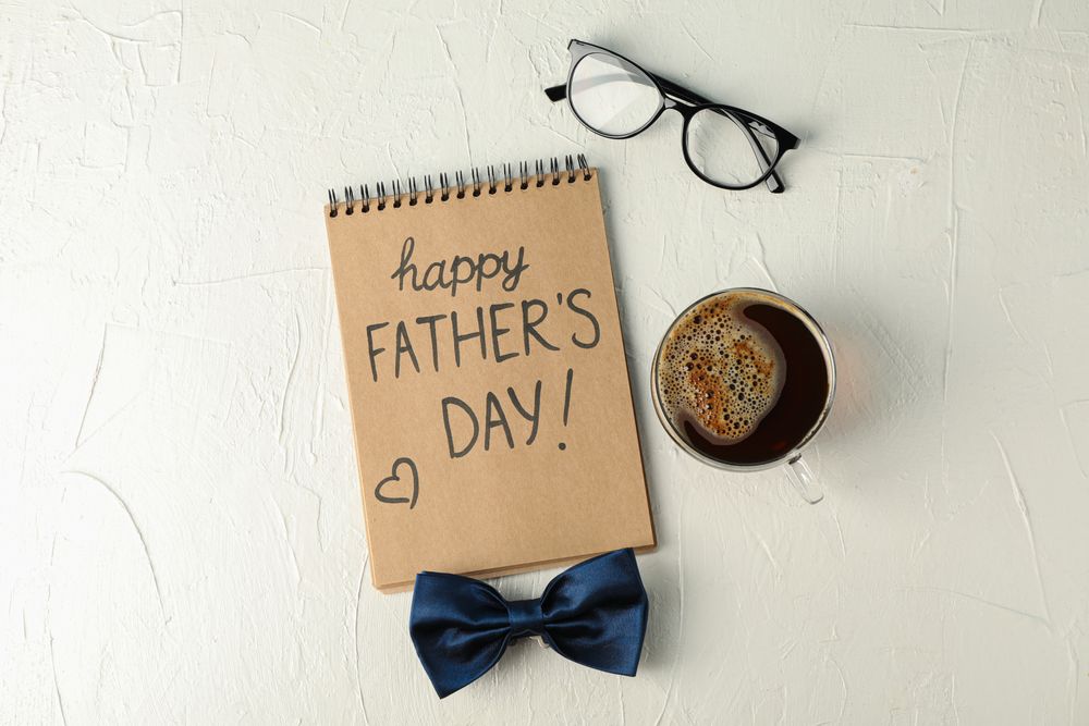 Notebook with inscription happy fathers day, blue bow tie, cup of coffee and glasses on plain background, space for text and top view