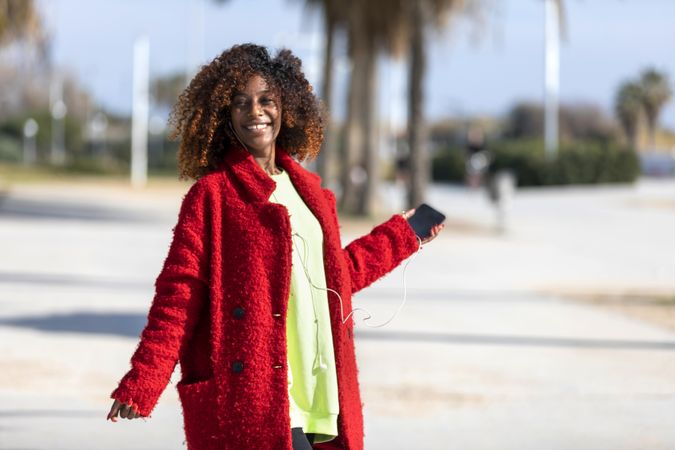Happy female in red jacket listening to music on phone standing on street with palm trees