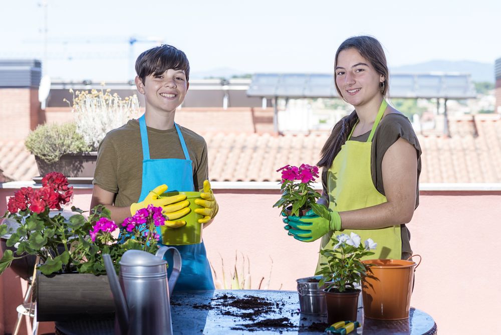 Two happy teenager wearing gardener apron posing at home terrace while holding a plant pot