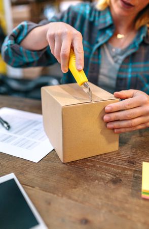 Unrecognizable woman with cutter unpacking order received by courier with merchandise