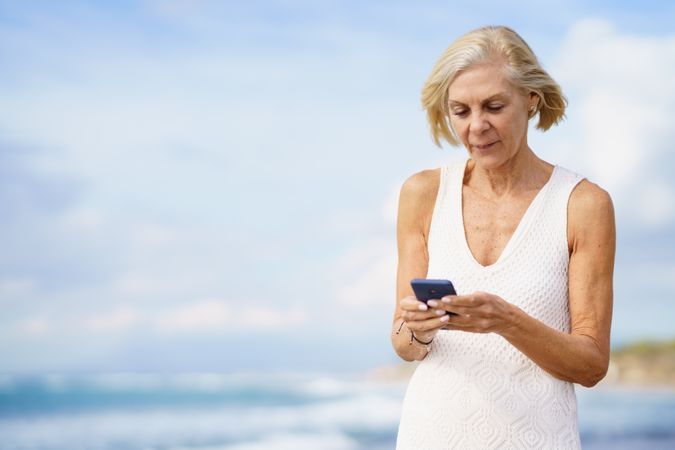 Older woman using her smart phone on the beach, copy space
