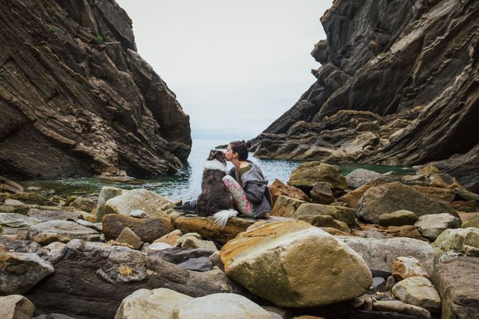 Traveling woman kissing dog sitting on boulder coast
