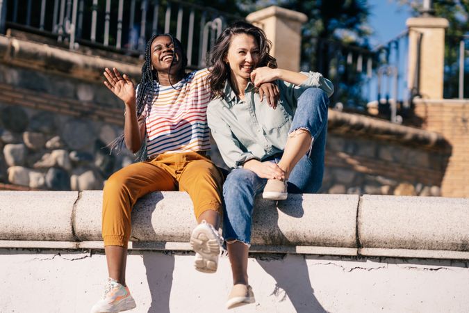 Close female friends laughing while sitting on outdoor wall on sunny day