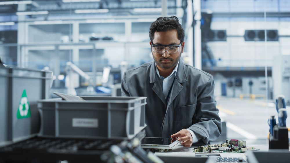 Indian Male Worker Disassembling Laptops To Recycle Electronic Components For Microchip Production At Automated Electronics Factory With Robotic Arms.