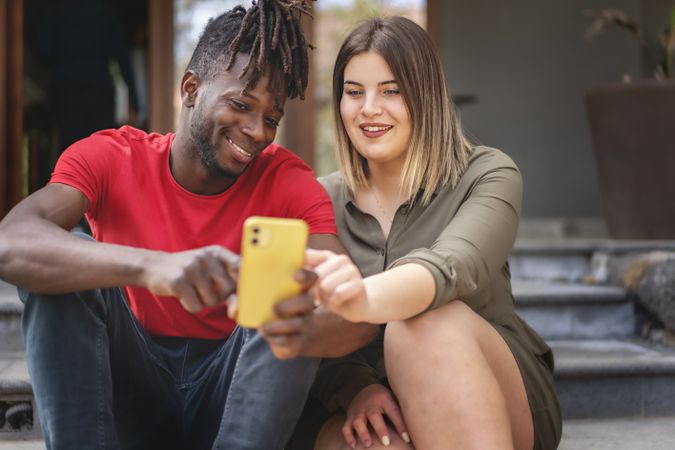 Two diverse friends, a Black man and white woman, sitting on outdoor stairs, looking at a smartphone content on a sunny summer day