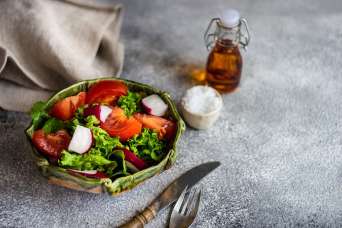 Bowl of salad with tomatoes, radish & lettuce on concrete background served with olive oil and salt