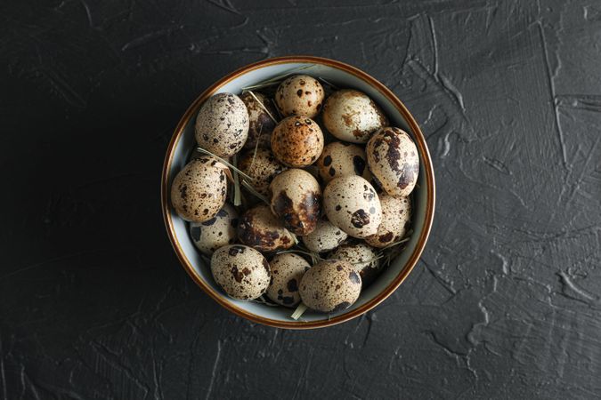 Many quail eggs in bowl on dark background, space for text and top view