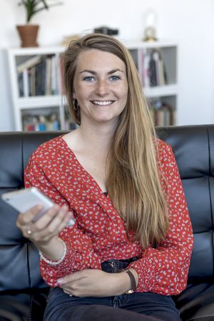 Young blonde woman sitting on sofa and holding mobile phone while smiling at camera