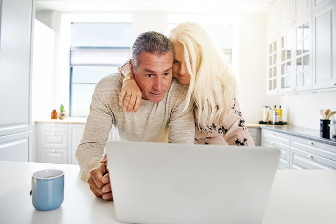 Woman draped over her husbands shoulder looking at laptop in kitchen