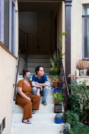 Loving couple sitting on stairs in front of apartment wearing masks and looking away