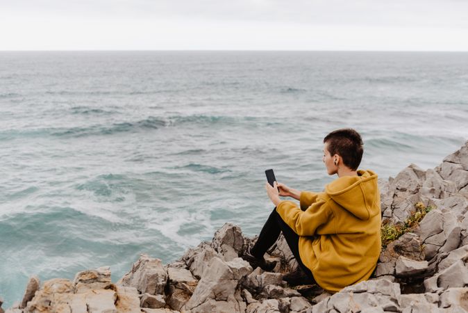Woman in yellow hoodie taking selfie on smartphone on rocky shore