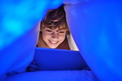 Joyful boy browsing tablet under blanket in late evening