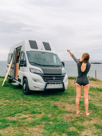 Female putting on wetsuit outside of motorhome parked on the coast, vertical