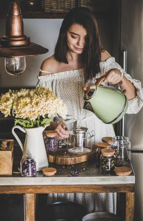 Woman in rustic kitchen pouring hot water from kettle into teapot
