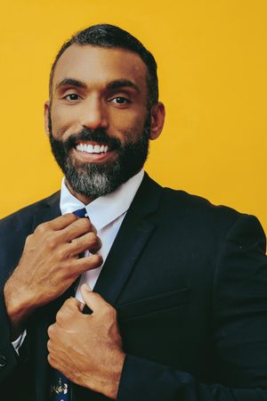 Smiling Black male in suit adjusting his floral tie in yellow studio