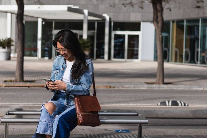 Long hair brunette Asian woman using mobile phone while sitting on a bench in the street