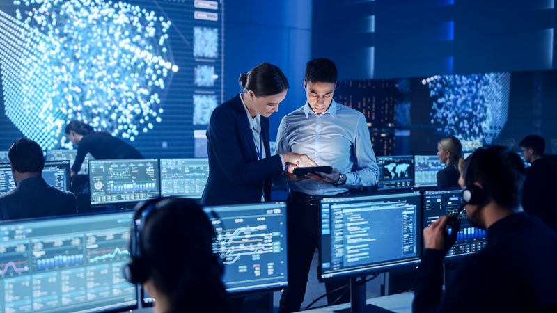 Executives Analyze Data on Tablet in Cybersecurity Operations Center. Team Monitors Global Network Activity on Multiple Screens.