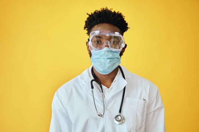 Portrait of male doctor in yellow studio with stethoscope, face mask and eye goggles