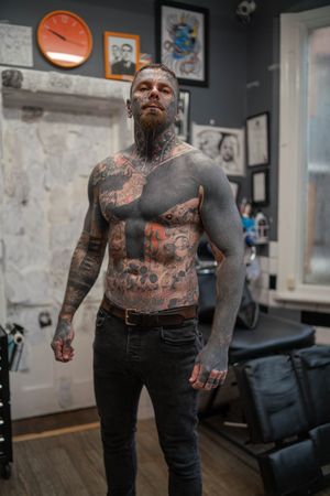 Shirtless muscular tough man with extreme full-body and face tattoos stands in tattoo parlor